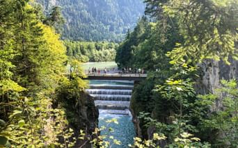 Lechfall waterfall near Füssen with turquoise water cascading over stepped weir. Visitors on footbridge, surrounded by lush forest and mountains.