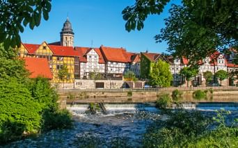 View of Hann. Münden old town with colorful half-timbered houses, red roofs, and church tower. River with weir in foreground, framed by green trees.