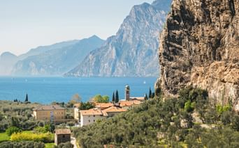 Torbole village with church tower nestled between dramatic cliffs and Lake Garda, surrounded by olive groves and Mediterranean vegetation.