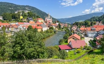 Malerischer Blick auf Murau mit rotgedeckten Häusern, Kirchturm und Fluss durch grünes Tal, umgeben von bewaldeten Bergen.