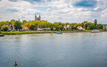 Der Rhein fließt an Speyer vorbei, mit den Doppeltürmen des historischen Doms über grünen Bäumen. Boote und Ufergebäude säumen das Ufer.