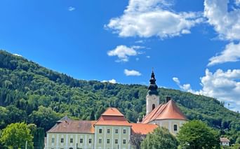 Stift Engelszell monastery with yellow buildings, red roofs, and church tower against forested hills under blue sky with white clouds.