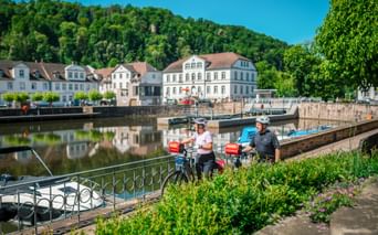 Zwei Radfahrer mit Tourenrädern am Barockhafen in Bad Karlshafen. Weiße historische Gebäude säumen das Ufer mit bewaldeten Hügeln dahinter.