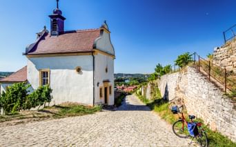 Weiße Kapelle mit orangefarbenem Dach und Glockenturm neben einem Kopfsteinpflasterweg. Ein Reiserad mit blauen Taschen steht an einer Steinmauer.