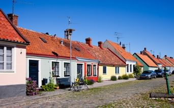 Reihe bunter einstöckiger Häuser mit orangefarbenen Ziegeldächern entlang einer Kopfsteinpflasterstraße in Simrishamn, Schweden, unter blauem Himmel.