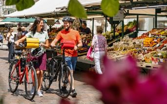 Two cyclists with e-bikes at a farmers market in Bozen, South Tyrol. Colorful fruit and vegetable stalls line the street under white awnings.