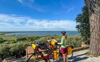 Female cyclist with touring bike and yellow panniers overlooking Lake Massaciuccoli. Wide lake view with green vegetation and blue sky.