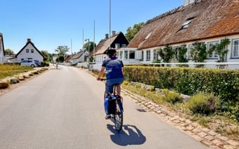 Cyclist riding through a quiet Danish village street on Zealand with traditional thatched-roof houses and hedges under a clear blue sky.