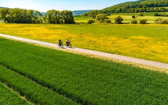Two cyclists on a paved path crossing green and yellow meadows in the Weserbergland region, with forested hills in the background.
