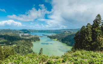 Panoramablick auf einen Vulkankratersee auf den Azoren mit türkis-grünem Wasser, umgeben von bewaldeten Hügeln unter blauem Himmel.