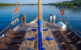 Sun deck of MS Macek with brown loungers and blue chairs. Passengers stand at the railing viewing the calm blue water and green hills.