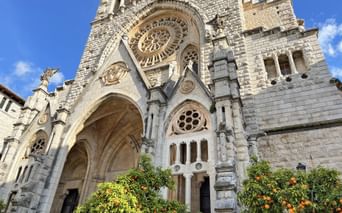 Gotische Kathedralenfassade in Sóller mit Rosettenfenster, Spitzbogeneingang und Orangenbäumen im Vordergrund vor blauem Himmel.