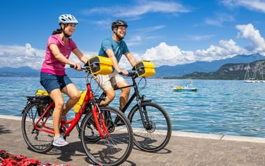 Zwei Radfahrer mit Helmen und gelben Taschen auf roten Rädern am Gardasee-Ufer in Bardolino, mit Blumen, blauem Wasser und Bergen.