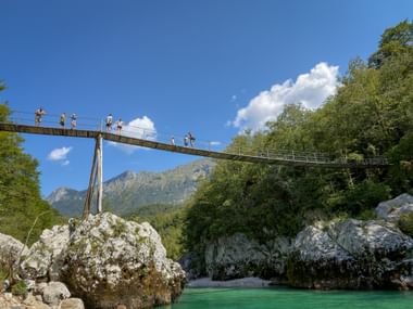 Suspension bridge near Kobarid spanning turquoise river between white rocks. People crossing bridge with mountains and blue sky in background.