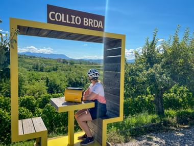 Radfahrer rastet an gelbem Aussichtspunkt mit Aufschrift Collio Brda, mit Blick auf Weinberge und Berge in Friaul unter blauem Himmel.