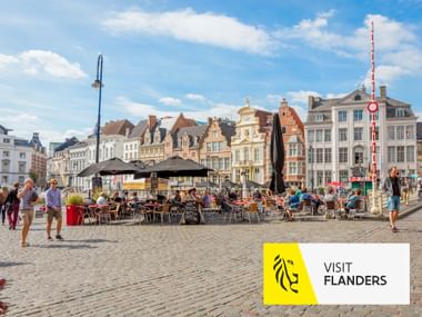 Kopfsteinpflasterplatz in Gent mit Café-Terrassen und historischen flämischen Giebelhäusern unter blauem Himmel. Visit Flanders Logo sichtbar.