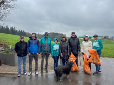 Gruppe von acht Personen und einem schwarzen Hund auf einem Weg mit orangen Müllsäcken. Grüne Felder und bewölkter Himmel im Hintergrund.