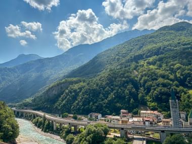 Dogna village nestled in a green Alpine valley with a stone viaduct bridge, turquoise river, and forested mountains under blue sky with clouds.