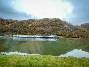 Weißes und blaues Flusskreuzfahrtschiff auf ruhigen Donaugewässern mit herbstlich gefärbten Hügeln im Hintergrund unter bewölktem Himmel.