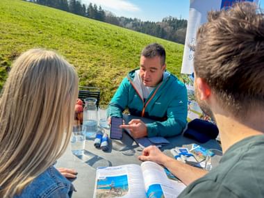 Three people at outdoor table reviewing Eurobike cycling brochures. Staff member in turquoise jacket consults with two guests against green meadow backdrop.