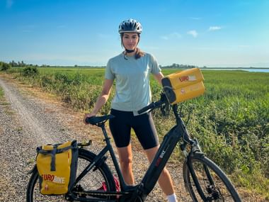 Radfahrerin mit Helm steht mit Fahrrad mit gelben Eurobike-Taschen auf Weg durch grünes Marschland bei Foci dello Stella.
