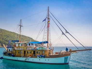 MS Panagiota, ein türkisfarbenes und gelbes Holzsegelschiff mit zwei Masten, ankert in ruhigem blauen Wasser nahe grünen Hügeln.