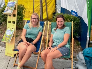 Two women in turquoise shirts sitting on deck chairs at the Trumer Triathlon event, with yellow and white banners in the background.