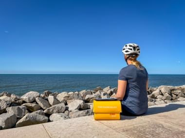 Radfahrerin mit weißem Helm sitzt an der Strandpromenade von Grado mit gelber Eurobike-Tasche, Blick auf die Adria und felsige Küste.