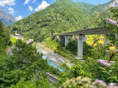 Concrete bridge spanning a turquoise river near Pontebba, surrounded by green mountains and forests along the Alpe Adria cycle route.