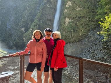Three cyclists on viewing platform at Liechtensteinklamm gorge with tall waterfall cascading down rocky cliff in background.