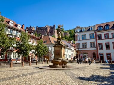 Kornmarkt in Heidelberg mit Madonna-Brunnen, bunten historischen Gebäuden und Heidelberger Schlossruine am Hang unter blauem Himmel.