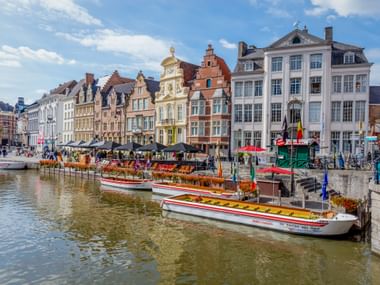Bunte historische Gebäude entlang des Graslei-Kanals in Gent mit Ausflugsbooten. Traditionelle flämische Architektur mit Treppengiebeln unter blauem Himmel.