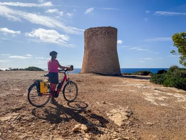 Radfahrerin in pinkem Oberteil mit rotem Fahrrad und gelben Packtaschen neben dem Wachturm Torre de Cala Pi an der Küste Mallorcas.