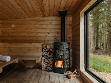 Interior of a wooden sauna with tiered benches, a black wood-burning stove with visible flames, stacked firewood, and a large window overlooking a forest.
