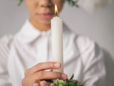 Person in weißer Kleidung hält eine brennende weiße Kerze mit grünem Laub beim Luciafest in nordischer Tradition.