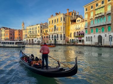 Gondolier in striped shirt rowing a black gondola with passengers on Venice's Grand Canal, with colorful historic palazzos lining the waterway.