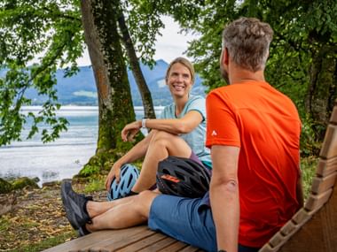 Two cyclists sitting on a wooden bench by Lake Tegernsee in Bavaria, with helmets beside them, surrounded by trees and mountain views.