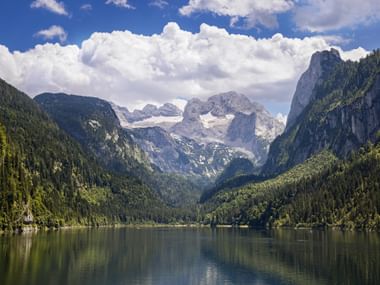 Gosausee im Salzkammergut mit ruhigem Wasser, das bewaldete Berge spiegelt. Dachsteinmassiv mit schneebedeckten Gipfeln im Hintergrund.