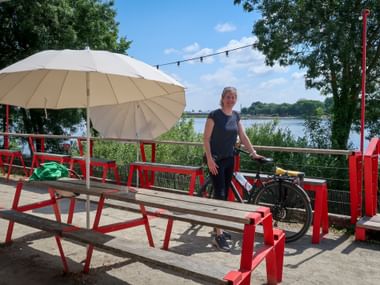 Female cyclist with bike standing at red picnic tables with umbrella on Loire riverside terrace near Nantes, with river and trees in background.