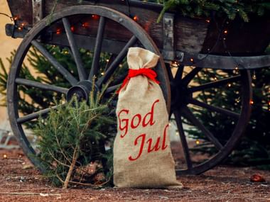 Beiger Jutesack mit roter 'God Jul'-Aufschrift und Schleife neben kleinem Tannenbaum vor rustikalem hölzernem Wagenrad.