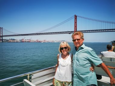 Two people on a boat on the Tejo River with the red 25 de Abril Bridge spanning across the water under a clear blue sky in Lisbon.