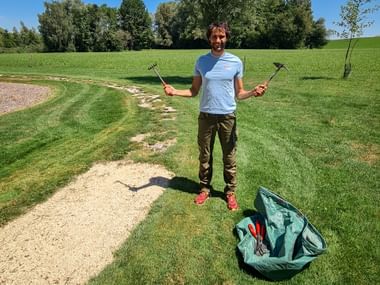 Smiling person in blue shirt holding garden tools in both hands, standing on a grassy path with a green bag nearby under clear blue sky.
