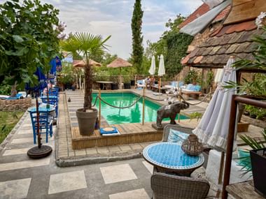 Außenterrasse eines Cafés mit türkisfarbenem Pool, Palmen, blauen Stühlen und Elefantenstatue. Steingebäude mit Ziegeldach im Hintergrund.