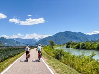 Two cyclists riding on paved Etsch cycle path alongside river with mountains in background under blue sky near Meran.