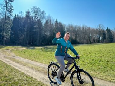 Man in turquoise jacket waving while cycling on gravel path through green meadow with forest and blue sky in background.