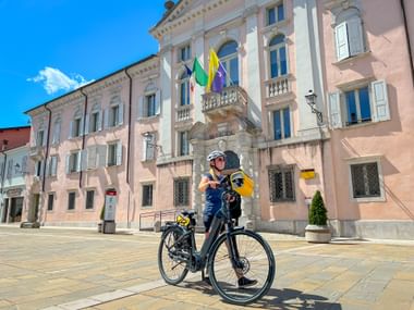 Radfahrer mit Kindersitz auf Stadtplatz vor Palazzo Torriani in Gradisca d'Isonzo. Historisches rosa Gebäude mit Fahnen unter blauem Himmel.