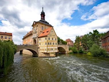 Historisches Fachwerkrathaus von Bamberg auf einer Steinbrücke über dem Fluss, mit orangefarbenem Dach und Turm unter bewölktem Himmel.