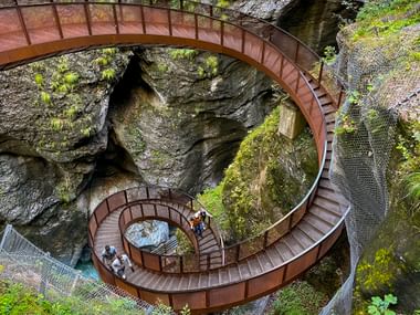Helix-shaped metal staircase winding through Liechtensteinklamm gorge with moss-covered rock walls and visitors below.