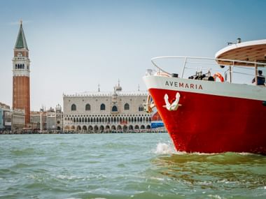Red and white ship Ave Maria sailing in Venice lagoon with St. Mark's Campanile and Doge's Palace visible in the background under clear blue sky.