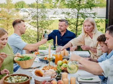 Familie versammelt sich um einen Tisch im Freien mit Essen und Getränken. Erwachsene und Kinder genießen ein Sommeressen mit Salaten und Brot.